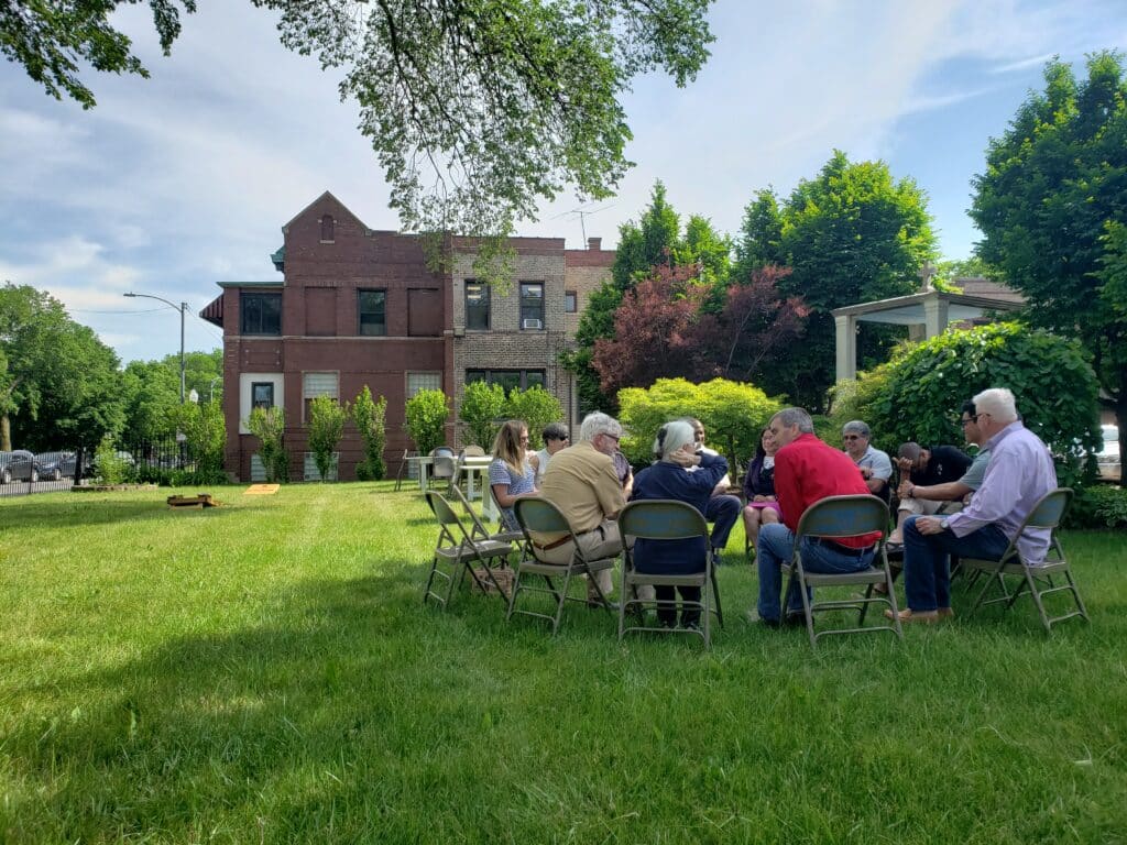 A group of people sit in a circle on folding chairs on a lawn in front of a house.