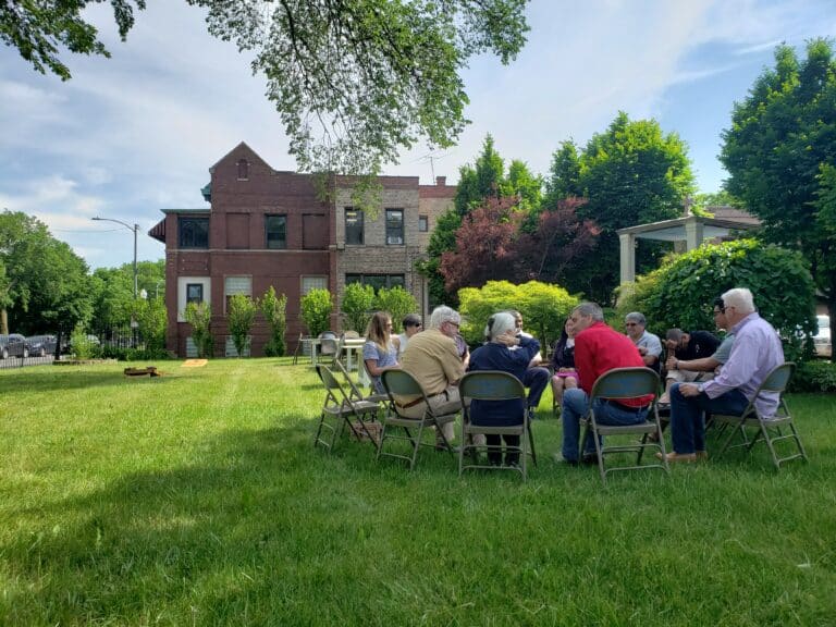 A group of people sit in a circle on folding chairs on a lawn in front of a house.