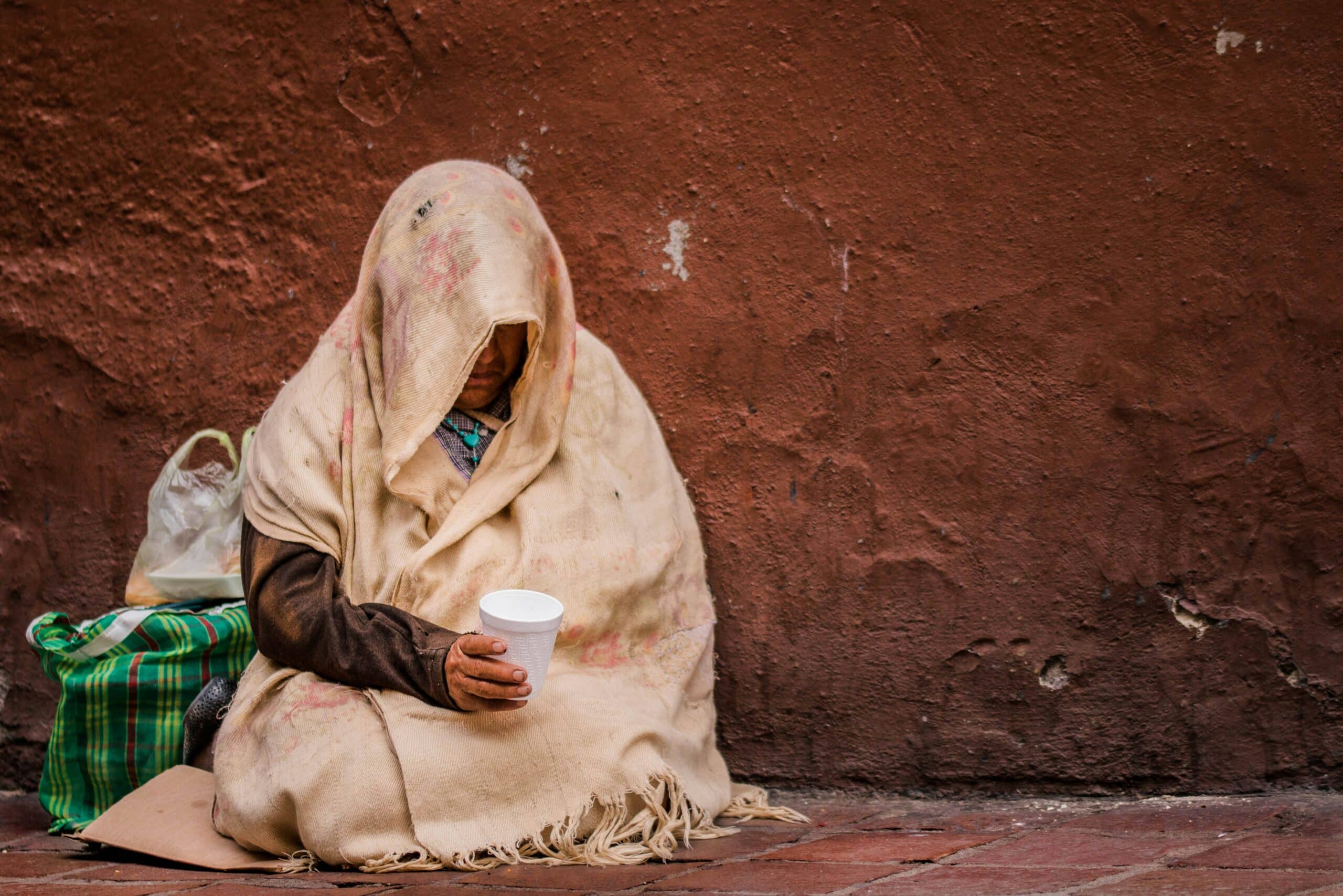 Person hidden by hood sitting against wall holding a styrofoam cup