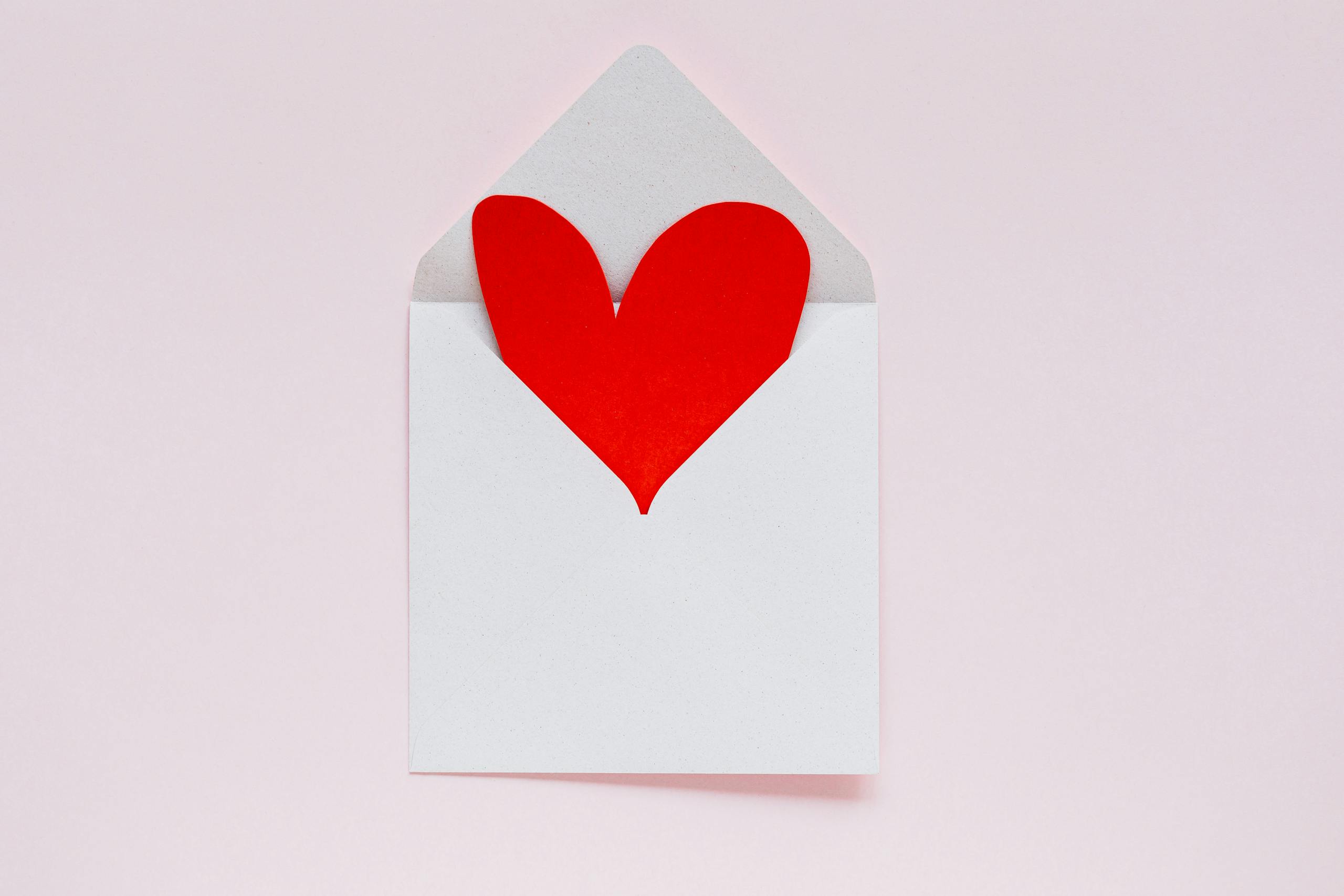 A red heart paper cutout in an envelope on a pastel pink background, ideal for Valentine themes.