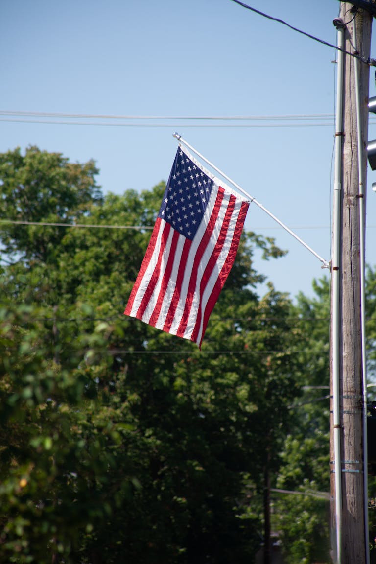 Patriotic American flag on a pole with trees and a clear blue sky background.